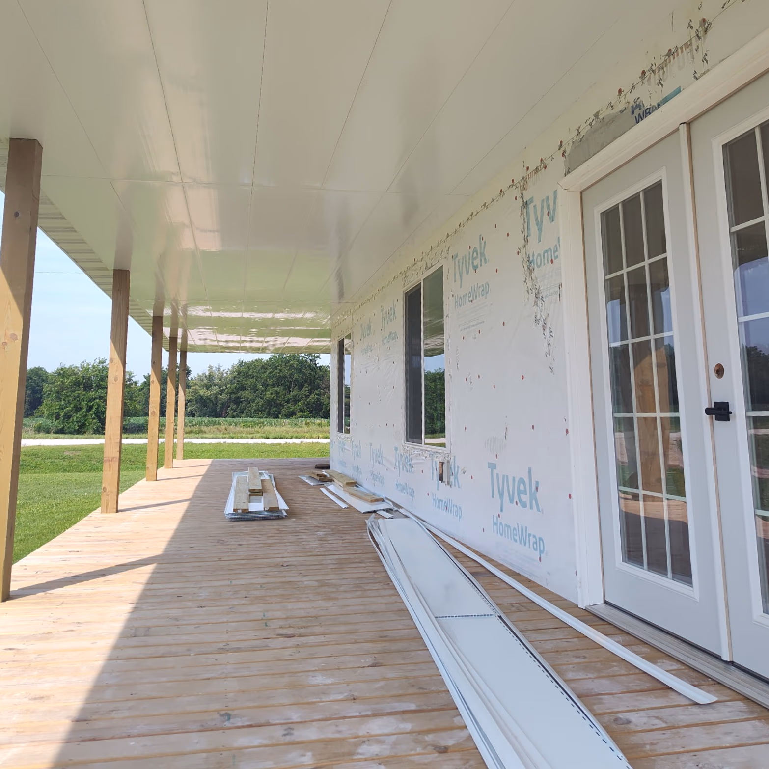 Wooden porch with posts, construction materials, and white wrapped house exterior with glass double doors.