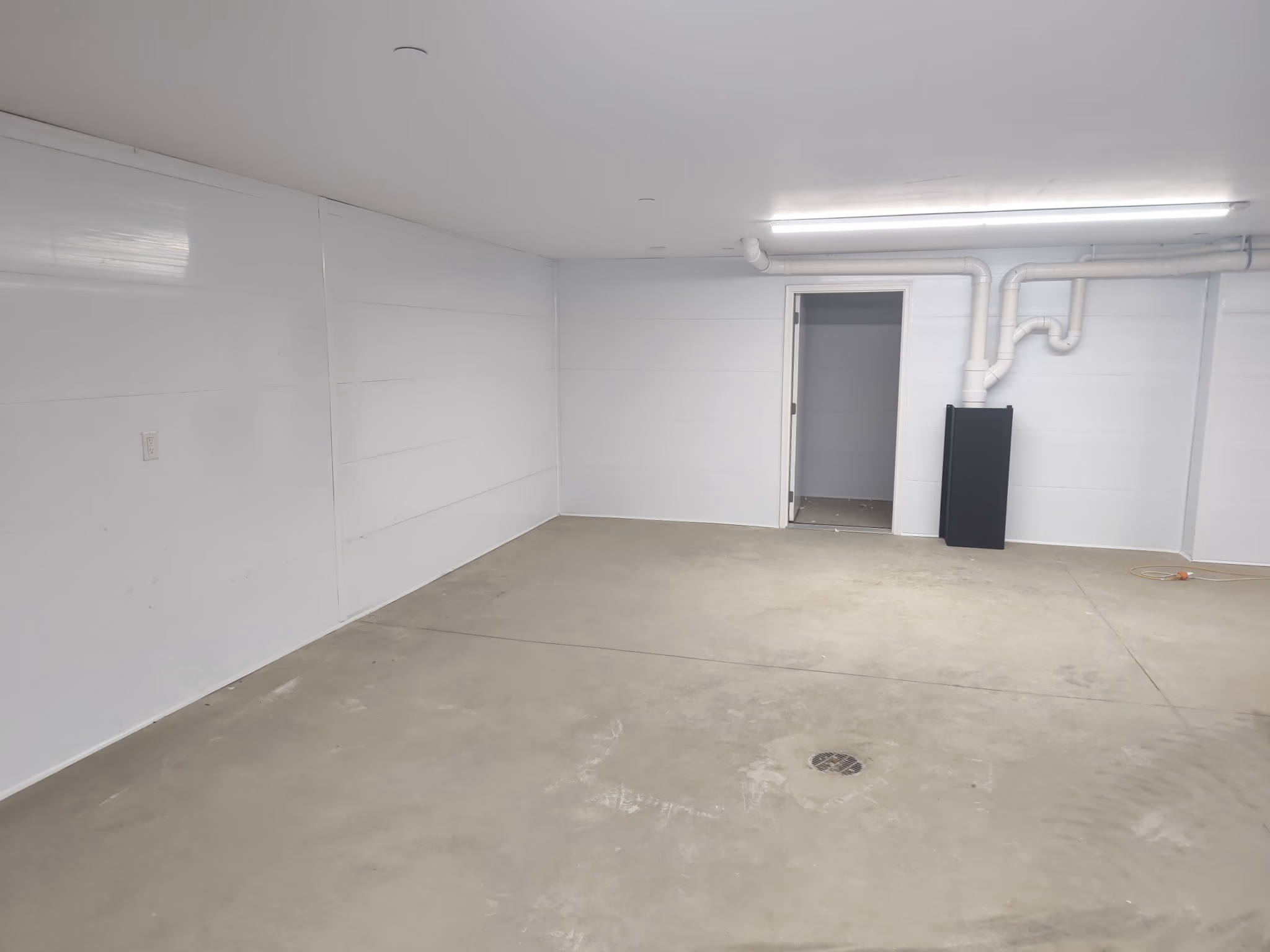 Empty basement room with white walls, a concrete floor, open door, and exposed white pipes on wall.
