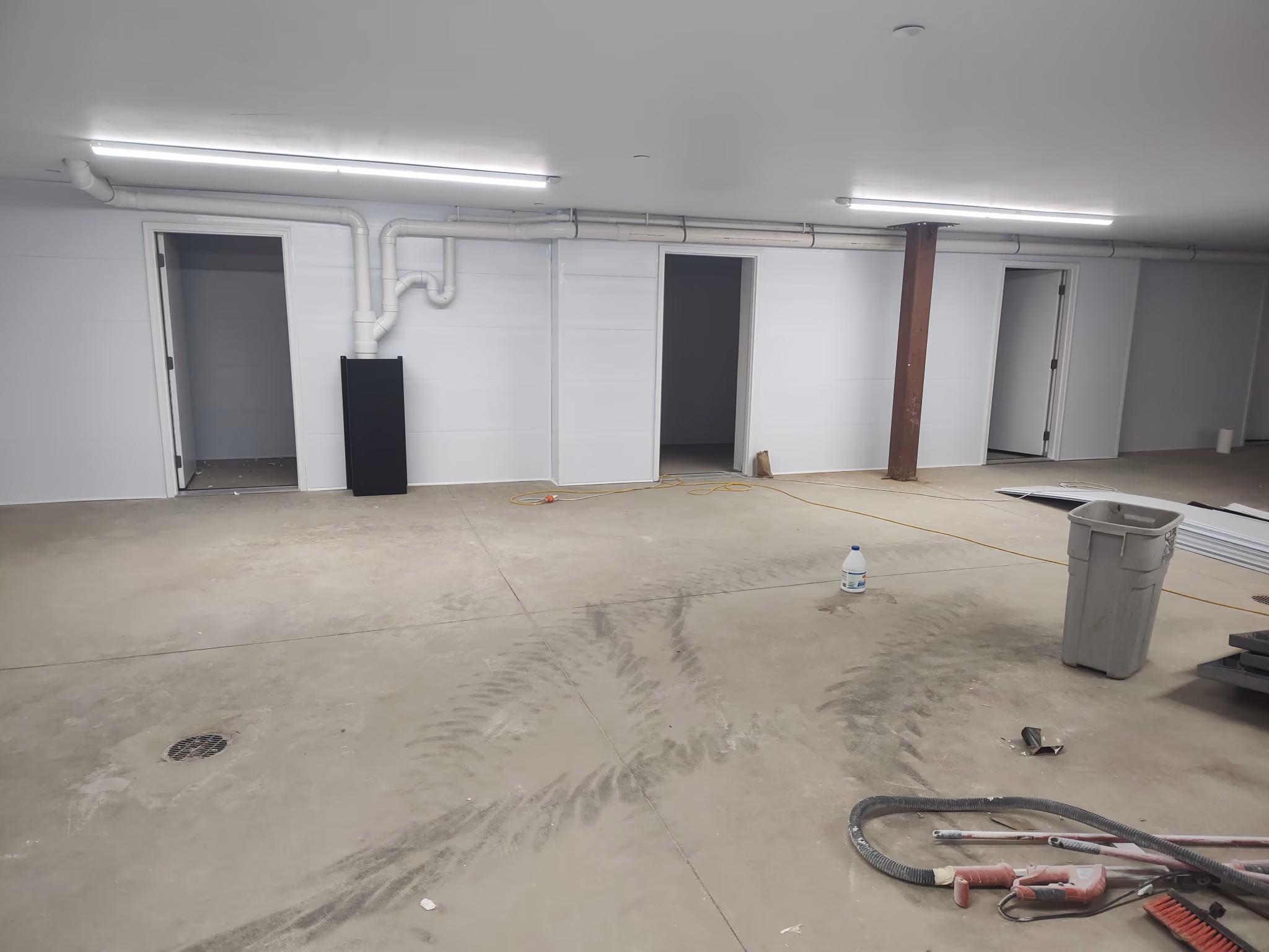 Unfinished basement with concrete floor, three open doorways, exposed pipes, tools, and trash bin.