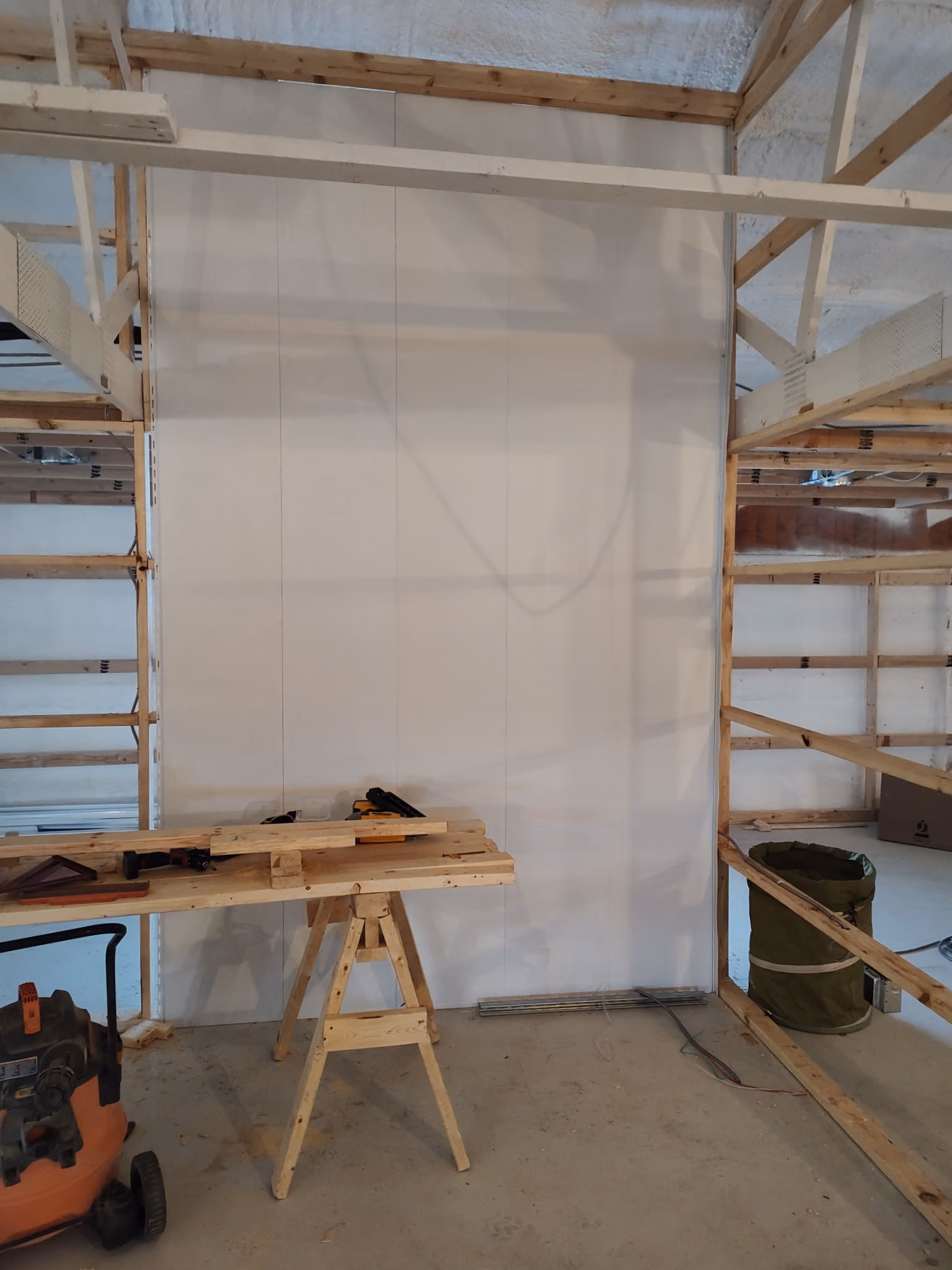 Construction site with wooden frame, sawhorse table, tools, and air compressor on concrete floor.