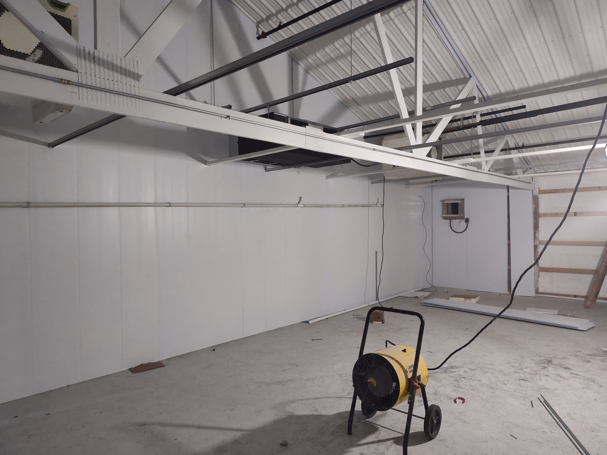 Empty industrial room with white walls, ceiling beams, and a yellow portable heater on wheels.