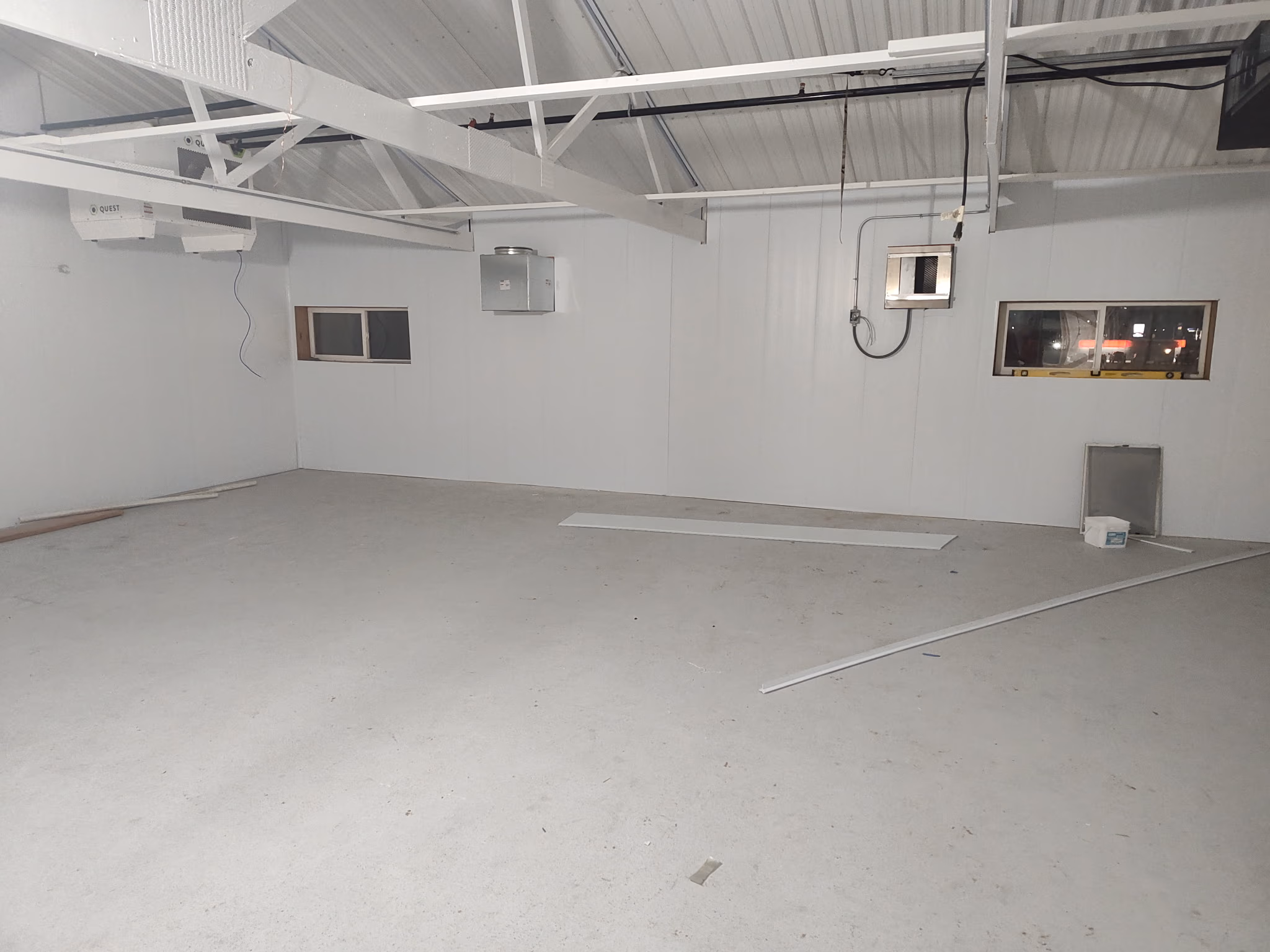Empty room with white walls, ceiling beams, two small windows, and industrial vents and wiring.
