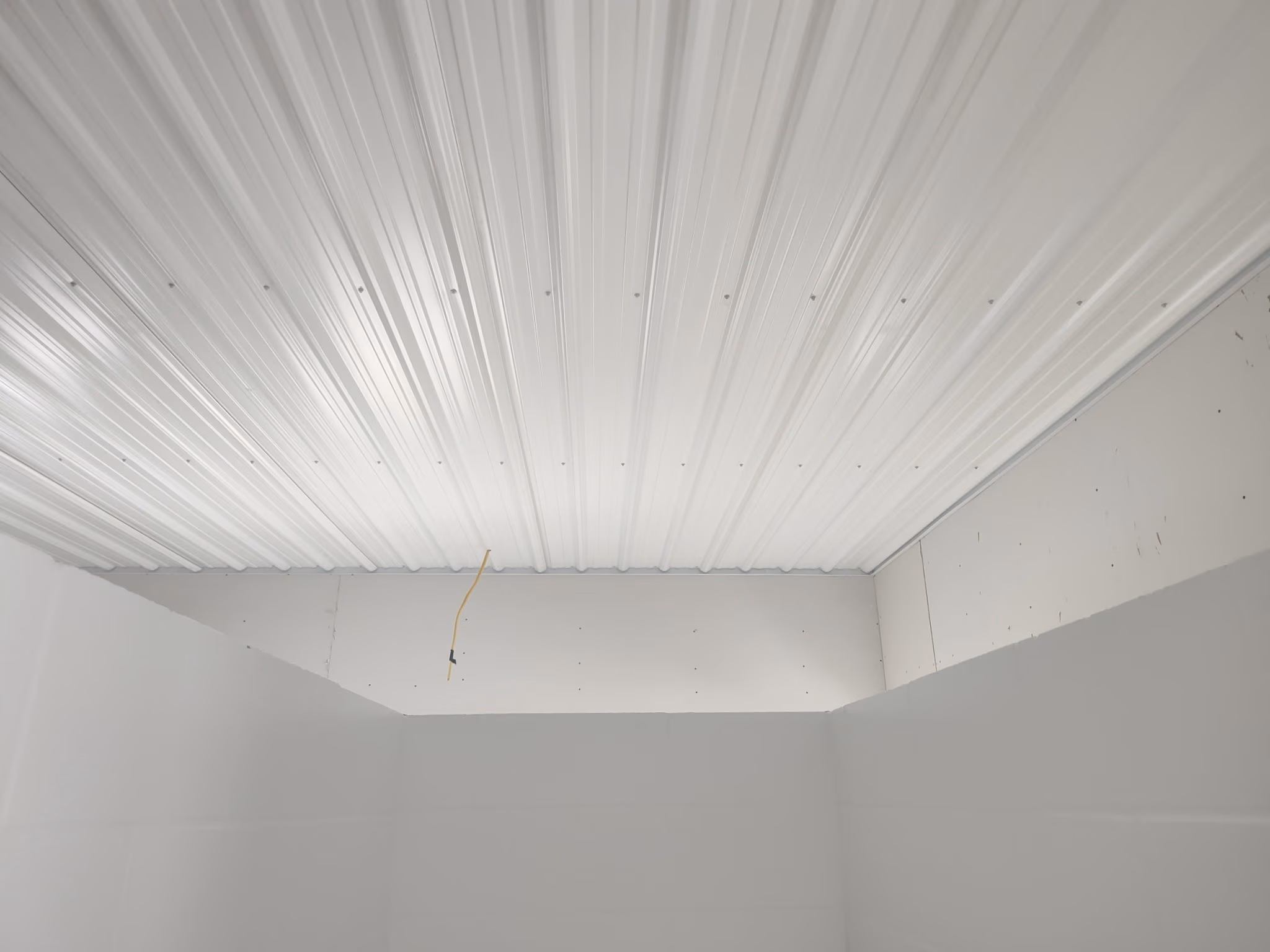 Unfinished room with white corrugated metal ceiling and bare drywall walls, yellow wire hanging down.