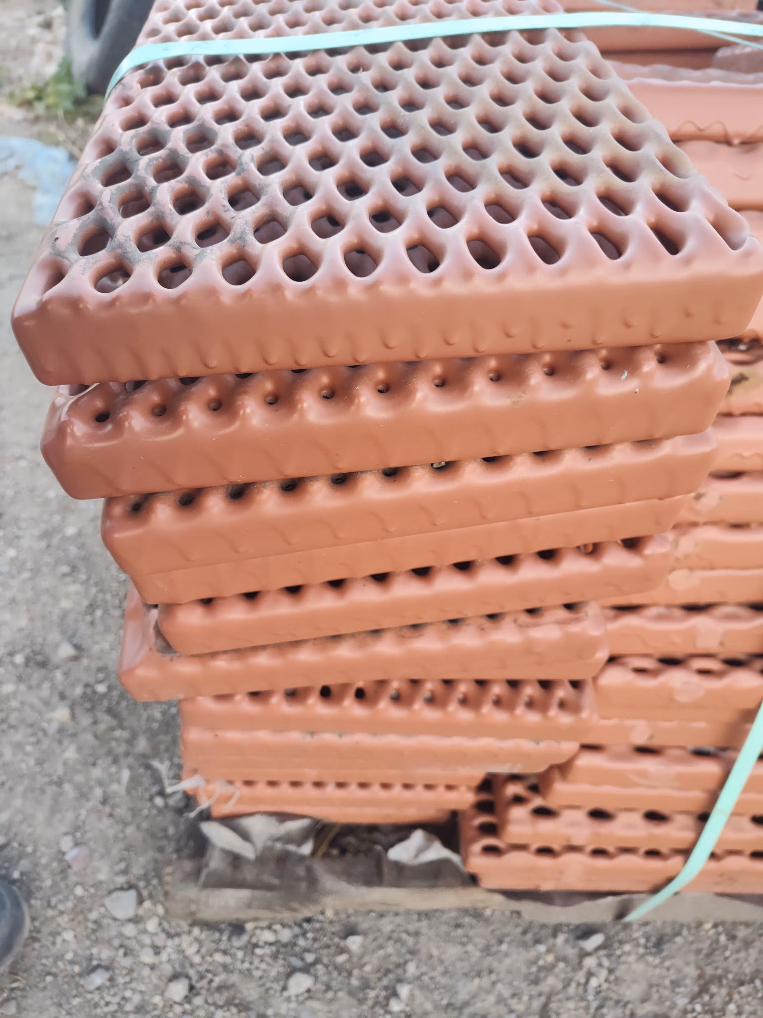 Stack of reddish-brown perforated mats bound with a green strap on a gravel surface.
