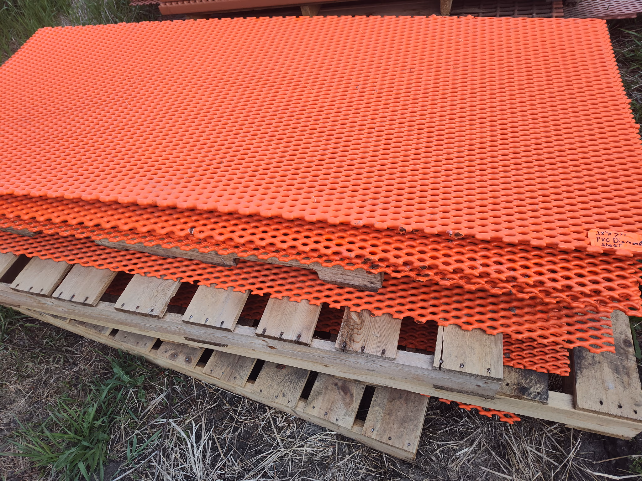 Stack of bright orange perforated plastic sheets on wooden pallets outdoors on grass and dirt.