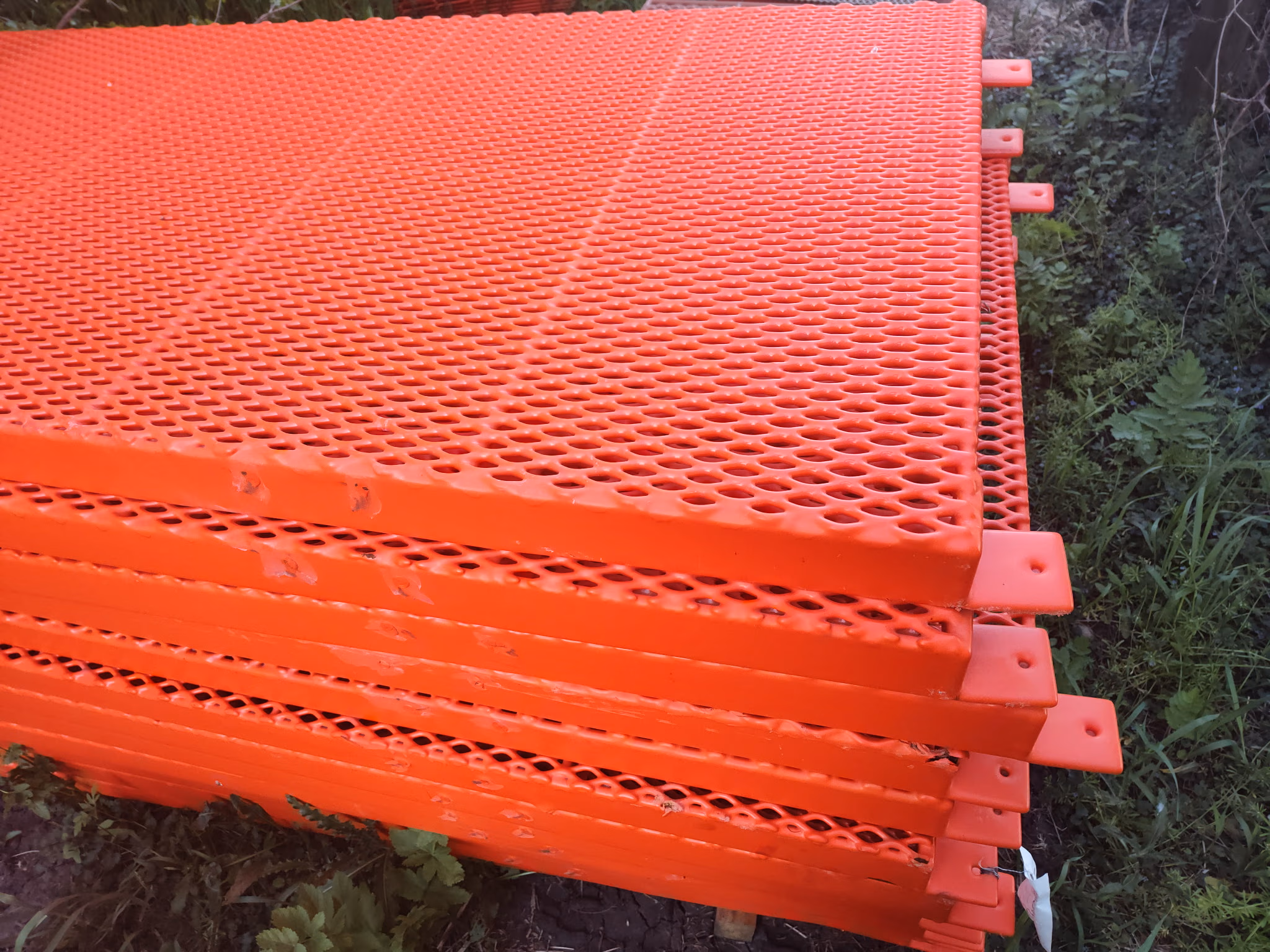 Stack of orange perforated metal panels resting outdoors on grass and plants.