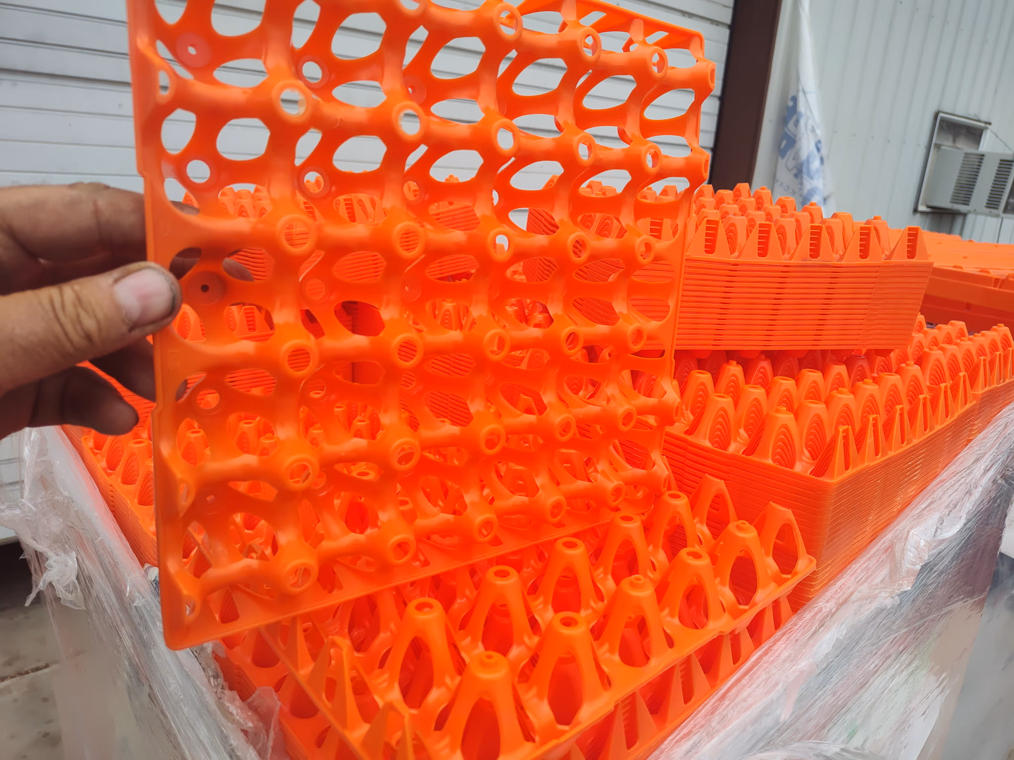 Hand holding a bright orange plastic grid panel among stacks of similar orange plastic grid pieces.