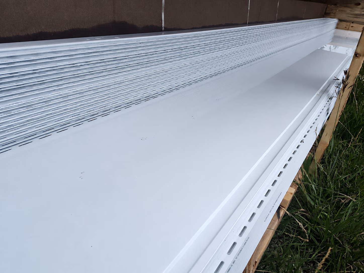 Stack of white metal siding panels with perforations, resting on wooden supports outdoors.
