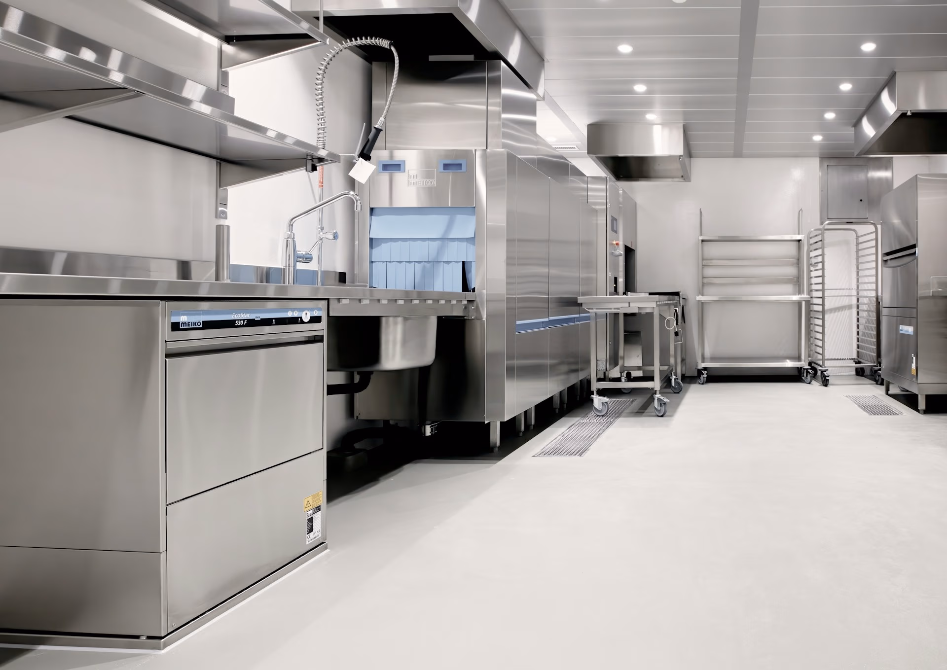 Modern industrial kitchen with stainless steel appliances, sinks, racks, and bright overhead lighting.